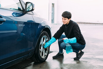Young woman cleaning the tire of a blue car using a sponge and a spray while wearing blue rubber...