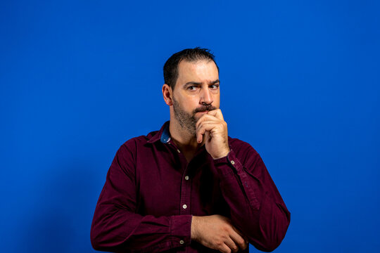 Portrait of young thoughtful man with hand near face with pensative expression against blue background in studio