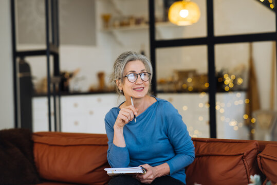Happy senior woman in glasses thinking and writes in a diary home