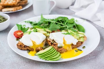 Healthy breakfast. Poached eggs on toast with avocado pieces, arugula, mizuna and chard leaves and cherry tomatoes on a plate on a served table. Flexetarian diet