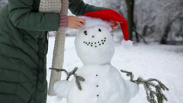 Woman puts festive red Santa hat on snowman in snowy winter park. Christmas celebration. Outdoor activities
