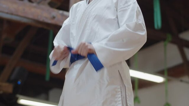 Medium shot of woman tying blue belt on kimono in practice room. Side view of female fighter preparing for training in gym, having morning karate workout. martial arts, sport concept