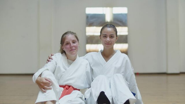 Front view of happy girls in kimonos hugging and sitting in gym. Medium shot of cheerful and beautiful athletes sitting on floor after karate training, posing for camera. Martial arts, sport concept