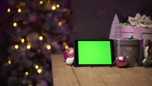Appearing on the right New Year's composition with a tablet green screen for mock-up, a horse, round gift boxes, balls on a wooden table. Lights twinkle on the Christmas tree.