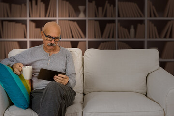 Fototapeta premium Senior Male Reading in a Digital Tablet Sitting at the Sofa at the House Living Room