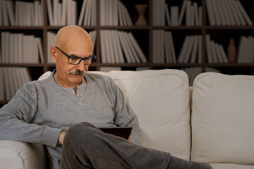 Fototapeta premium Senior Male Reading in a Digital Tablet Sitting at the Sofa at the House Living Room
