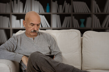 Fototapeta premium Senior Male Reading in a Digital Tablet Sitting at the Sofa at the House Living Room