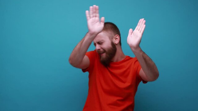 Positive full of energy bearded man dancing to the music, having fun, resting and enjoying life, party, entertainment, wearing orange T-shirt. Indoor studio shot isolated on blue background.
