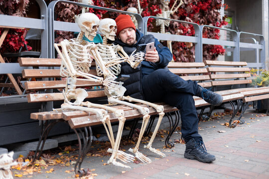 A man with a beard in a vest and a hat sits and looks at his phone on a bench with two skeletons, which also have phones. Day, cloudy, autumn.
