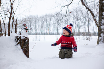 Fototapeta na wymiar little boy in a red Christmas sweater and a red hat stands in the snow and looks at the snowman. Winter outdoor activities.