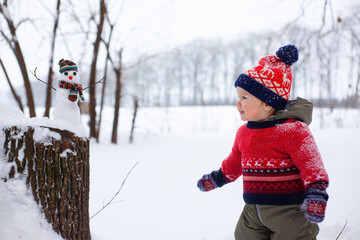 little boy in a red Christmas sweater and a red hat stands in the snow and looks at the snowman. Winter outdoor activities.