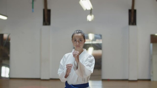 Front view of serious girl performing basic karate stances. Medium shot of concentrated teenager in kimono with blue belt exercising in gym, punching, lunging, looking at camera. Sport concept