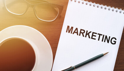Marketing word in notebook on the desk in sunlight next to glasses, a cup of coffee and a pen