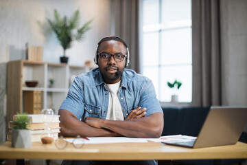 Young african man in headset and eyeglasses sitting at desk with laptop and papers and looking at camera. Male freelancer having meeting online while staying at home.