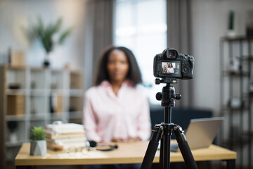 Blur background of pretty african american woman with dark hair sitting at desk and filming video blog. Focus on modern digital camera fixed on tripod.