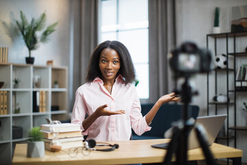 Charming african american woman in pink shirt talking and gesturing while recording video on modern camera. Female blogger sitting at home and doing live stream.