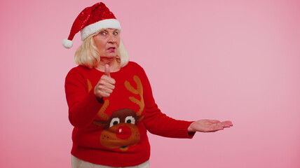 Elderly grandmother woman wears red New Year deer sweater showing thumbs up, pointing at right on blank space. Place for your advertisement logo on pink background studio. Happy Christmas holiday sale