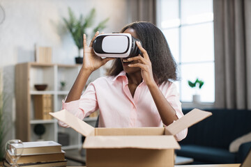 African american woman sitting at table with opened parcel box and testing new VR glasses. Happy female buyer unboxing package with internet order.