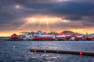 Naklejka premium Norwegian fishing village and sea coast at sunset. Lofoten islands, Norway. Winter landscape with red houses, blue water, dramatic cloudy sky with golden sun rays. Norwegian traditional red rorbu