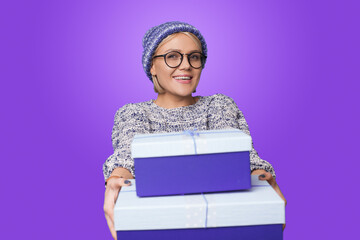 Caucasian woman wearing knitted hat giving Christmas presents at camera. Celebrating winter holidays. Posing on purple background.