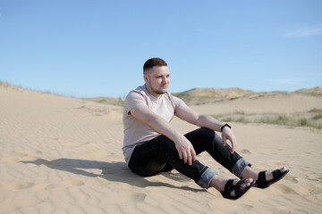 Fototapeta na wymiar Young tired man sitting on the top of sand dune in desert, sand is al around and some greens. Hot summer weather, concept of travelling around planet. Great Europen desert in Ukraine. Active lifestyle