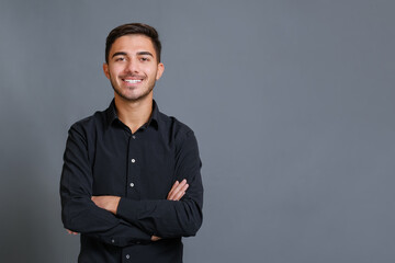 Handsome young unshaved man in black shirt with crossed arms looking at camera against gray wall and smiling. Satisfied caucasian guy looking at camera with a big laugh, copy space