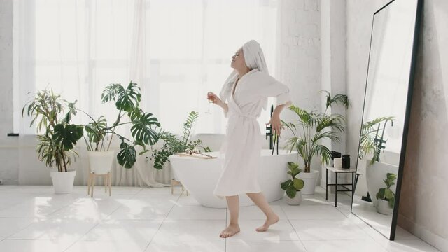 Young caucasian woman in white bathrobe and towel on her head dancing with glass of champagne after morning shower at home, slow motion. People, joy and music concept. Woman alone happiness concept