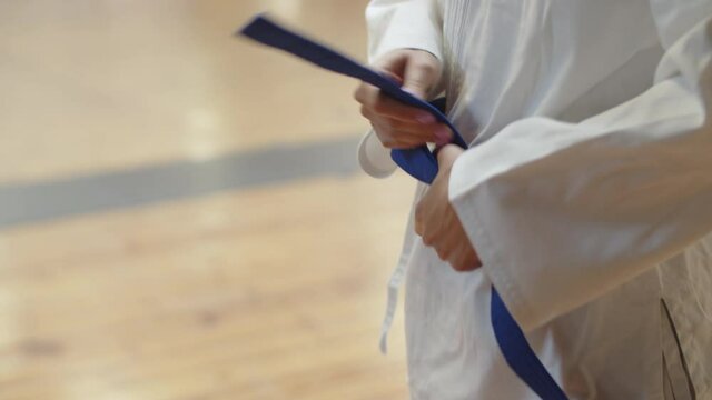 Close-up shot of woman standing in gym and tying belt on kimono. Side view of unrecognisable female fighter preparing for karate training in practice room. martial arts, sport, judo concept