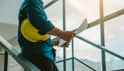 Architect man holding hardhat and blueprint while standing outdoors and against building structure.