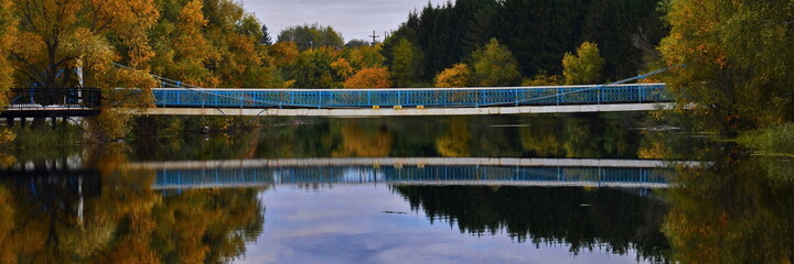 Panoramic view on long metal bridge across the river with reflection in water on the background of an autumn landscape
