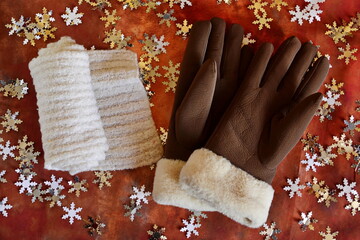 Woman winter clothing accessories. Brown soft leather gloves with fur trim and rolled white down knitted warm scarf on imitation fallen snowflake on brown background. Top view