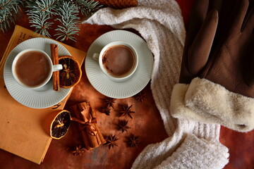 Winter drink. Pair cup of hot chocolate on book. Top view on brown table decorated with cinnamon sticks, star anise, slice of dry lemon, christmas tree twig, woman gloves and white down scarf