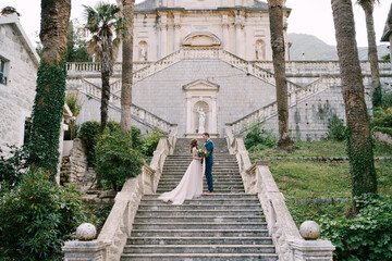 Bride and groom stand on the steps in the garden of the church in Prcanj. Montenegro - Powered by Adobe