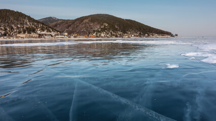 Intersecting cracks are visible on the blue shiny ice of the frozen lake. City houses on the shore. A wooded mountain range against an azure sky.  Reflection. Baikal