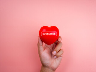 Subscribe concept. Word SUBSCRIBE on red heart ball in hand on pink pastel background with copy space, minimal style. Following social media, buying on the internet, ordering products online in apps.