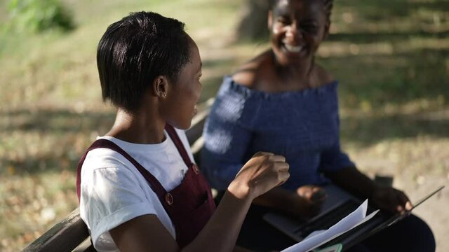 Smiling happy African American female student gesturing victory high-five with friend sitting in sunshine in summer park. Beautiful woman talking with groupmate rejoicing in slow motion outdoors