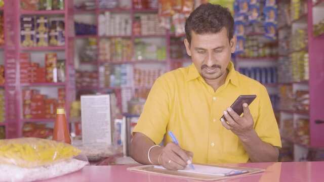 Indian groceries business man noting orders while talking with customer on mobile phone at kirana shop - concept of distance shopping, home delivery service.