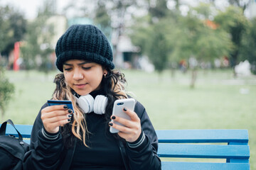 Latin teenager college girl sitting on a bench at the park buying online with credit card