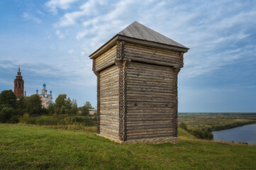 Log old watchtower on the high bank of the Kolva River (Russia, Cherdyn) on a green lawn on a sunny summer day. View of the ancient churches. Blue sky with white clouds. Rural pastoral. Bright photo 