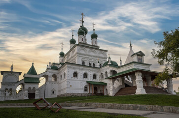 A view of an ancient temple with many green domes, carved stone elements, columns, arches and other architectural decorations on a summer morning. Yellow-blue cloudy sky. Russia, Solikamsk 