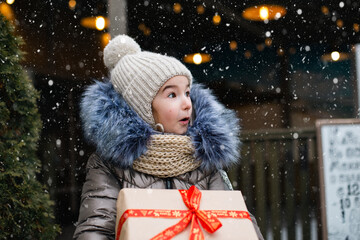 Portrait of surprised girl with a gift box for Christmas on a city street in winter with snow on a festive market with decorations and lights. Warm clothes, knitted hat, scarf and fur. Copy space