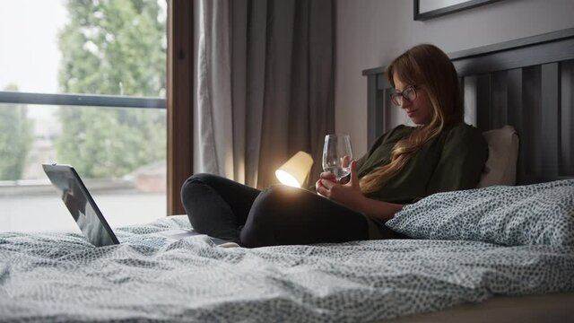 Charming woman sitting on the bed, drinking white wine and watching the laptop in the bedroom