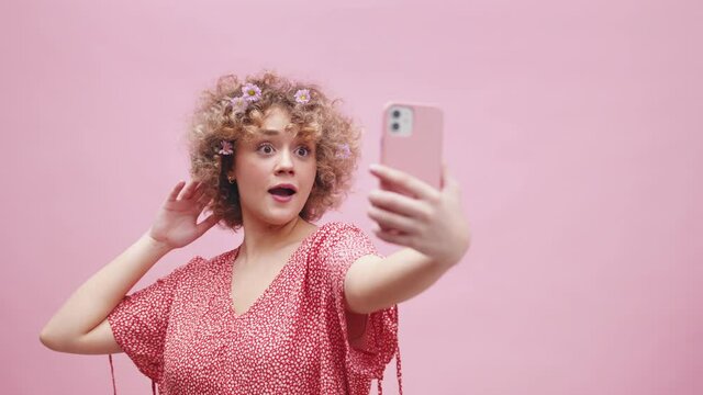 Young Woman With Flowers in a Curly Hair Posing For Selfie Camera of Smartphone. Studio Shot, Pink Background Full Frame