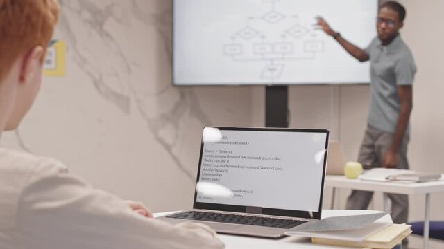 Over-shoulder view of boy sitting at desk in classroom, typing computer code on laptop, listening to lecture of blurred teacher