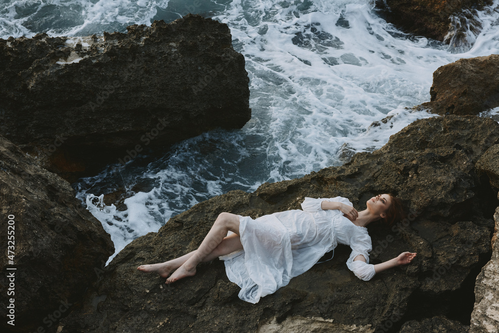 Sticker attractive woman with long hair in long white dress wet hair lying on a rocky cliff view from above