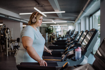 Mid adult overweight woman with headphones exercising on treadmill in gym - obrazy, fototapety, plakaty