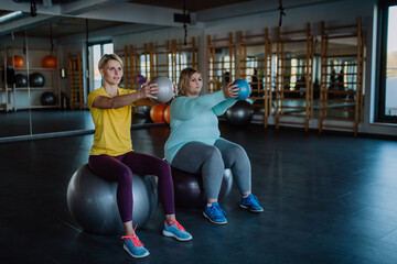 Fototapeta na wymiar Overweight woman with dumbbells sitting on fintess ball and exercising with personal trainer in gym