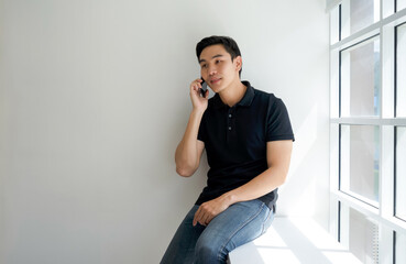 Young asian man in black collar t-shirt, sit and talking on mobile phone beside the window.