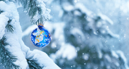 Blue glass ball with Christmas angel hanging on snowy fir tree, natural winter background. symbol...