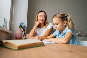 Low-angle side view of pretty little 6 years kid daughter writing with young mom tutor sitting at home table by window. Loving adult parent mother teaching school child girl helping with homework.